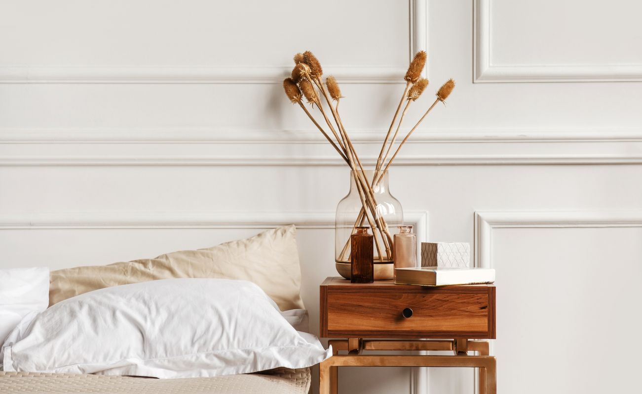 Ein Schlafzimmer mit einem Bett, einem Kissen, einem Nachttisch aus Holz und einer Vase mit Trockenblumen vor einer hellen Wand.